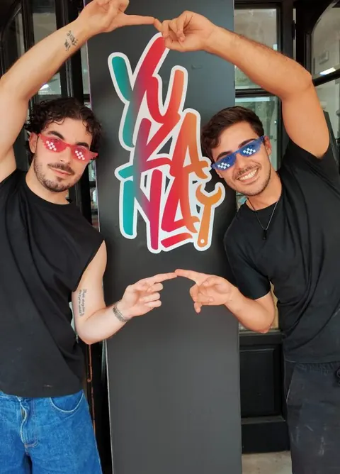 Los dos chicos de Yukana con gafas de colores, haciendo un corazón alrededor del logo